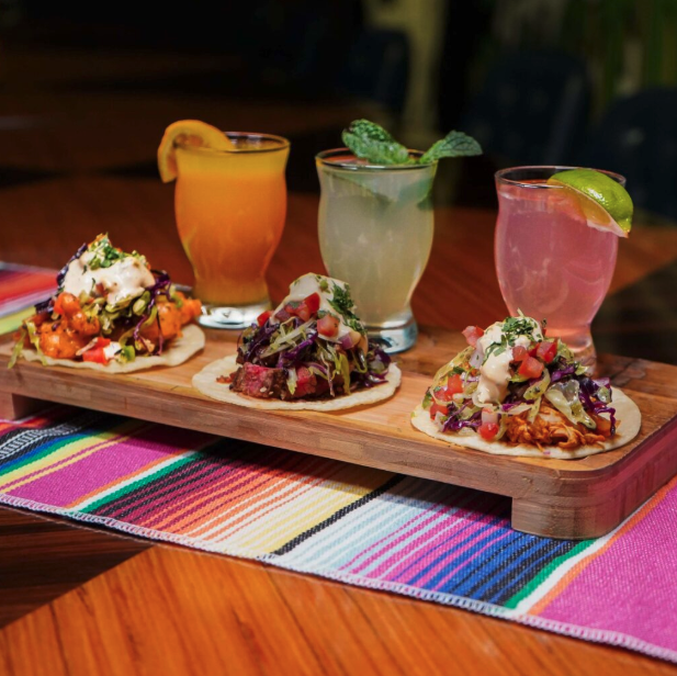 Three tacos with assorted toppings and three colorful cocktails served on a wooden board over a vibrant table runner.