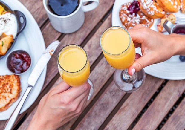 Two people clinking glasses of orange mimosas over a wooden table set with coffee, pastries, and breakfast dishes.