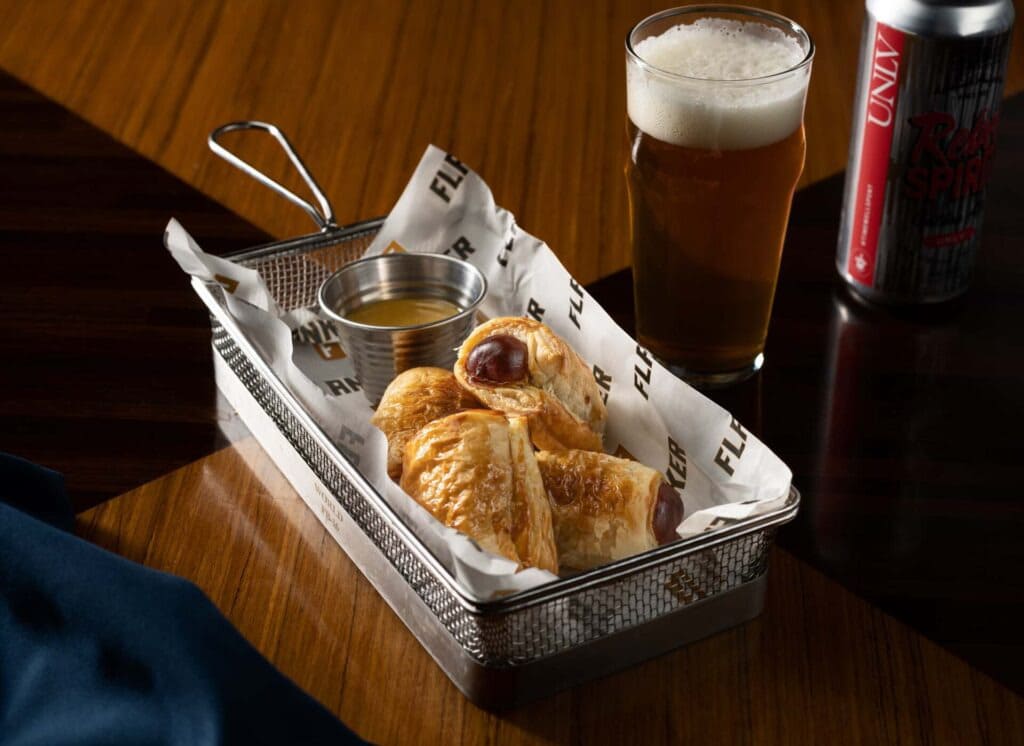 Basket of pigs in a blanket with dipping sauce, served with a pint of beer on a wooden table.