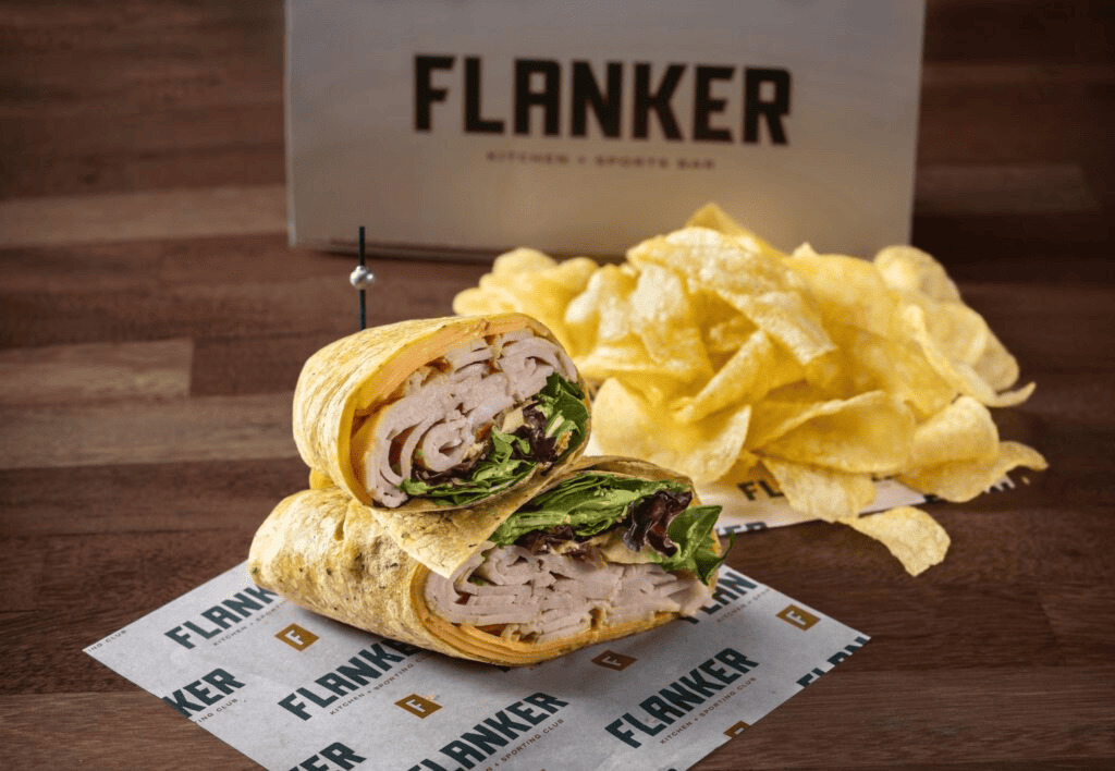 Turkey wrap sandwich cut in half, served with potato chips on branded Flanker paper.