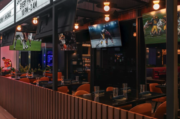 Indoor sports bar dining area with wooden panel dividers, orange chairs, and multiple screens showing various sports games.