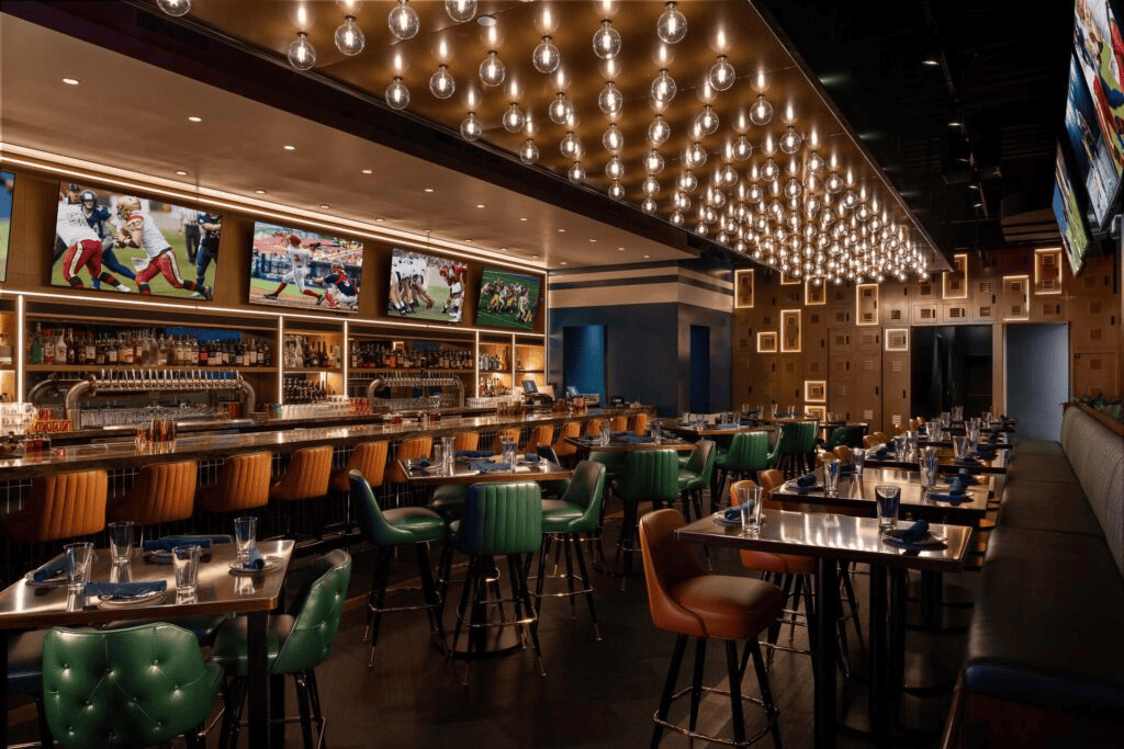 Modern sports bar interior with TVs displaying football games, high-top tables with place settings, and stylish lighting fixtures hanging from the ceiling.