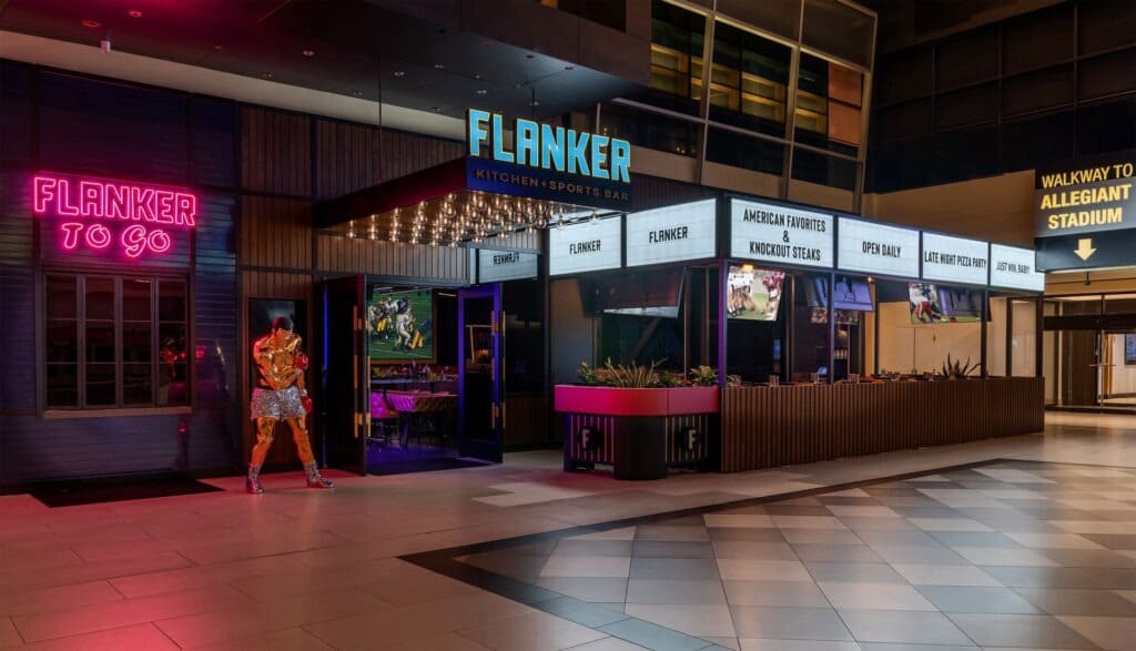 Front view of Flanker Kitchen + Sports Bar at night, featuring neon signage, marquee-style lighting, a metallic boxer statue, and a visible walkway sign to Allegiant Stadium.