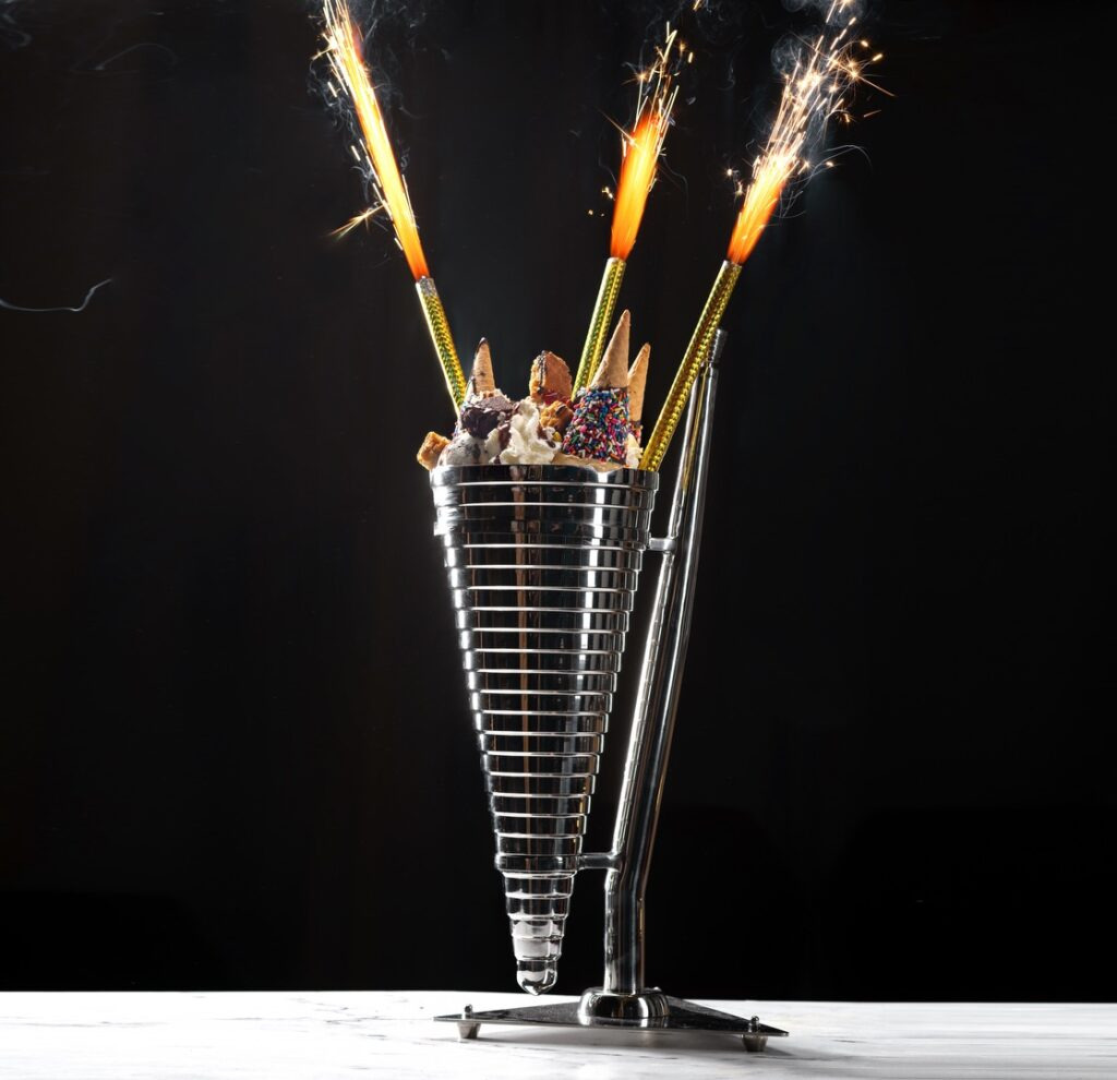 Cone-shaped metal dessert holder filled with sweet treats and topped with lit sparklers against a dark background.