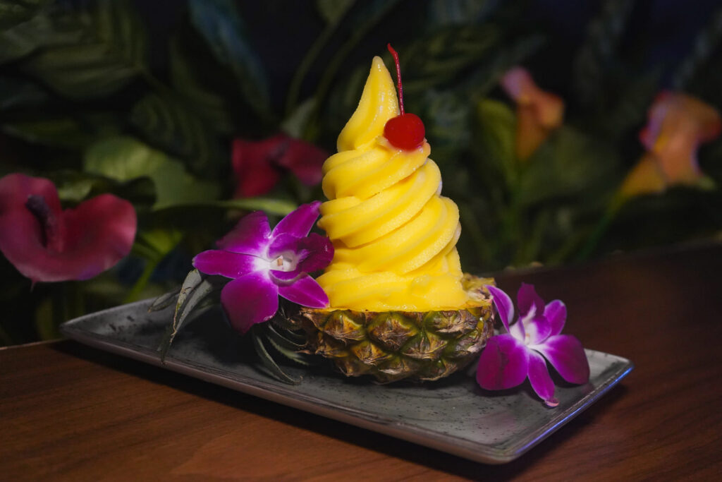 Image pop-up Pineapple soft serve dessert served in a pineapple shell and topped with a cherry and orchid flowers.