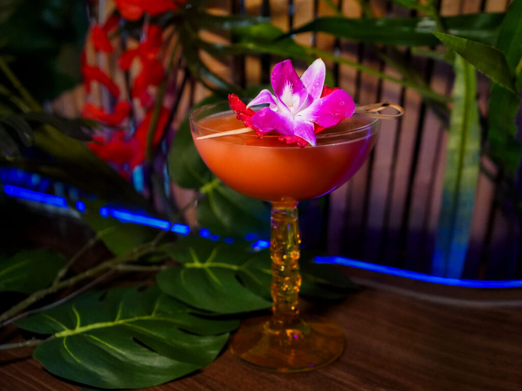 Image pop-up Tropical cocktail in a coupe glass garnished with a purple orchid and fruit, surrounded by lush tropical foliage.