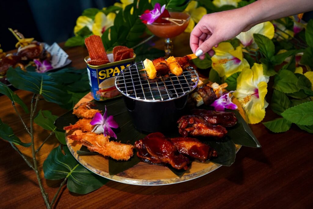 Image pop-up Tropical-inspired appetizer platter featuring grilled skewers, barbecue wings, coconut shrimp, and cocktails with floral garnishes.