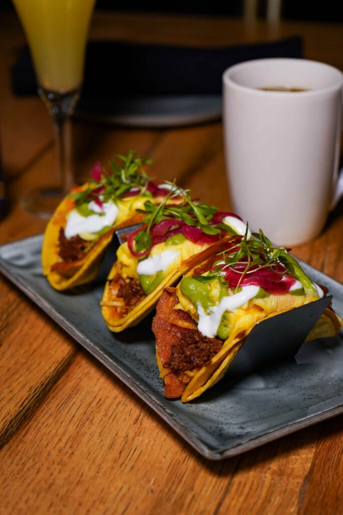 Image pop-up Three breakfast tacos filled with eggs and seasoned meat, topped with avocado sauce, crema, pickled red onions, and fresh greens.