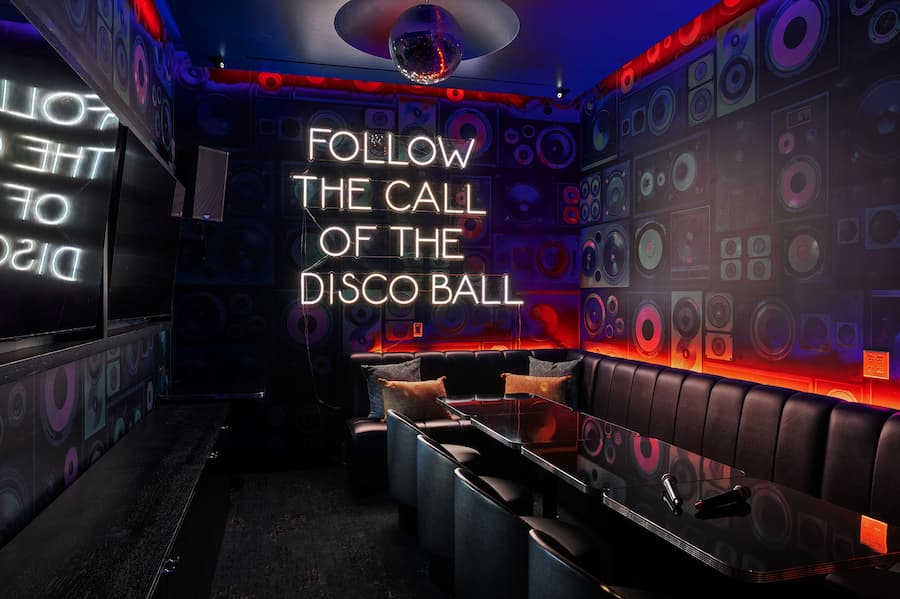 Private karaoke room with lounge seating, colorful speaker-patterned walls, a disco ball, and a neon sign reading “Follow the Call of the Disco Ball.”
