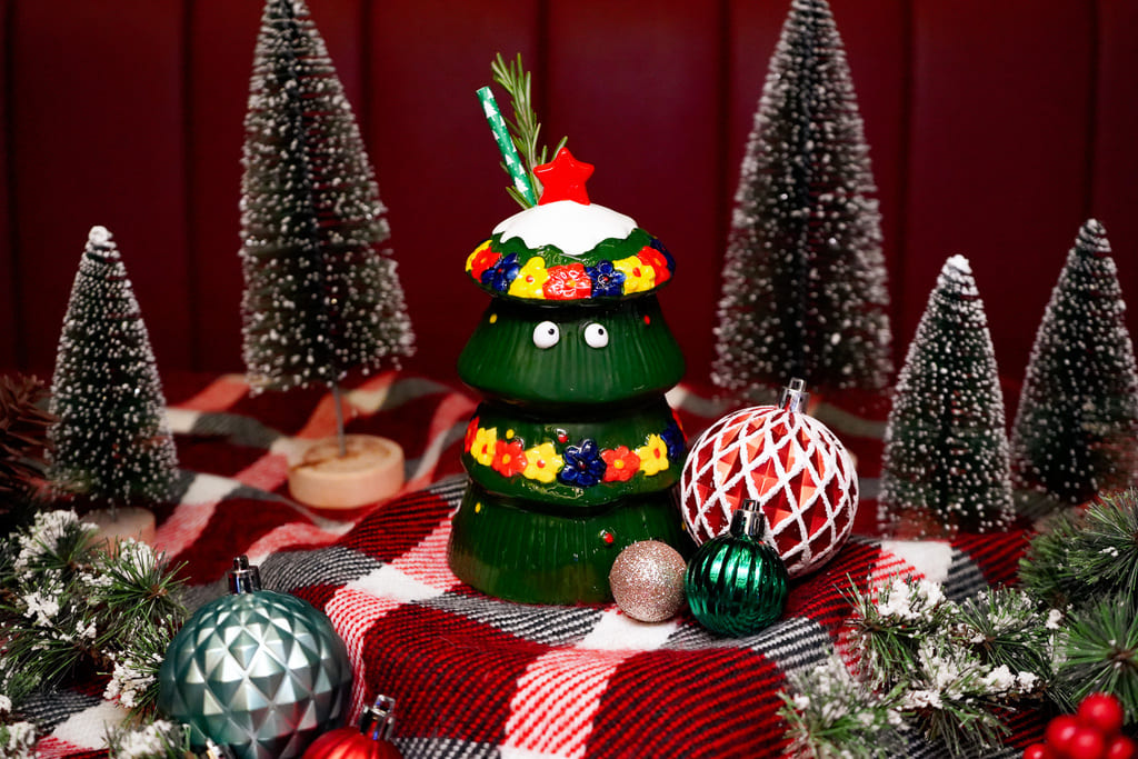 Image pop-up Green Christmas tree-shaped cocktail vessel decorated with colorful ornaments and a red star, surrounded by miniature snowy trees and Christmas baubles.