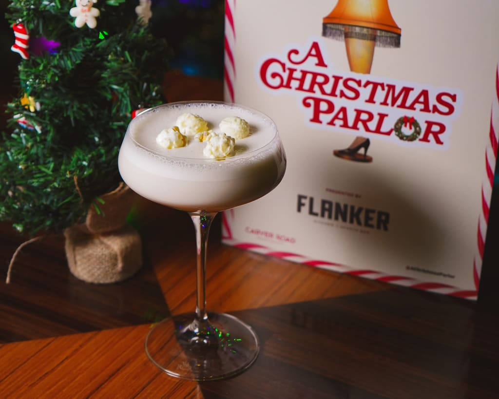 Image pop-up Creamy white cocktail garnished with small scoops of topping in a coupe glass, displayed beside a miniature Christmas tree and Flanker