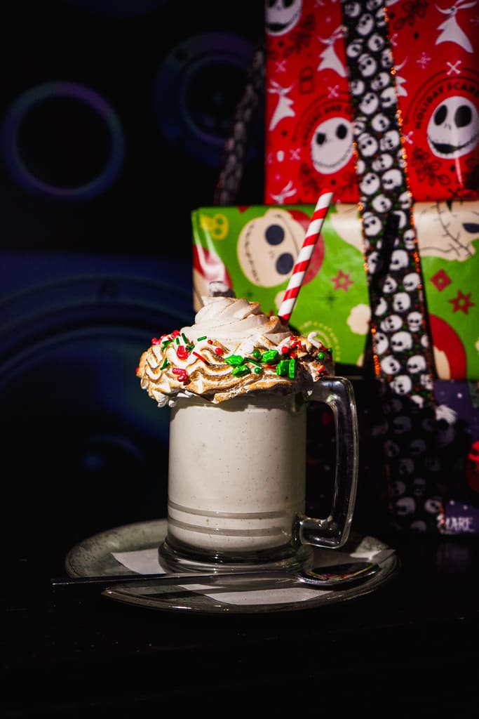 Image pop-up Creamy holiday milkshake topped with whipped cream, colorful Christmas sprinkles, and a red-and-white striped straw beside wrapped holiday gifts.