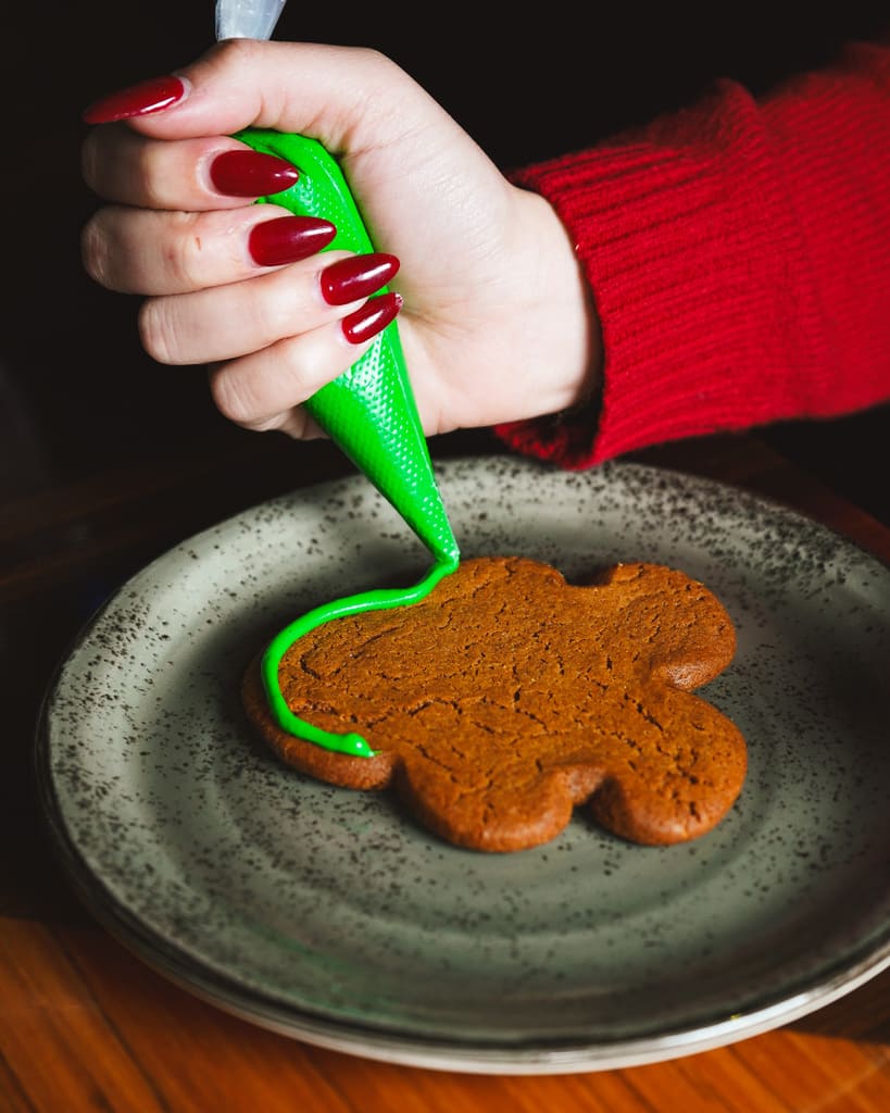 Image pop-up Hand with red-painted nails applying bright green icing to a gingerbread cookie on a ceramic plate.