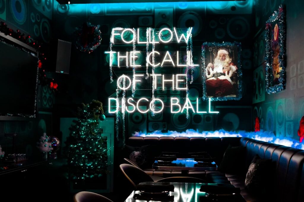 Image pop-up Festively decorated karaoke lounge with a glowing “Follow the Call of the Disco Ball” neon sign, Christmas trees, holiday garlands, and black leather seating.