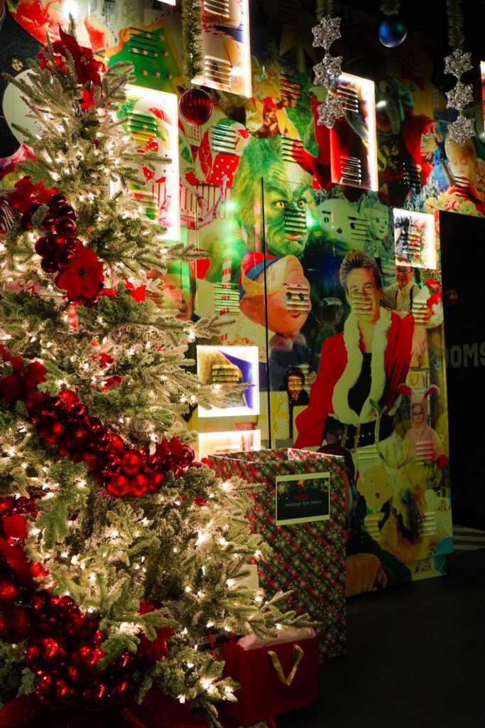 Image pop-up Snow-covered Christmas tree decorated with red ornaments and warm lights beside a holiday toy drive donation box and colorful Christmas movie mural.
