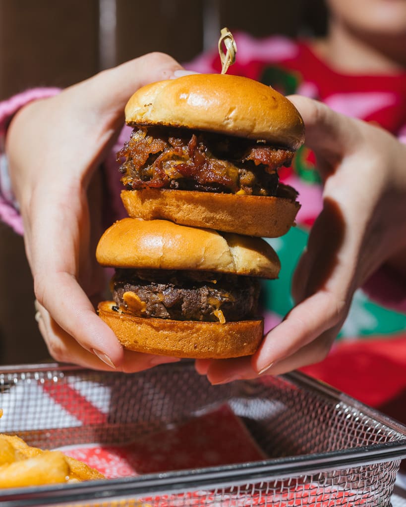 Image pop-up Two mini beef burgers stacked vertically with golden brioche buns, held between two hands above a metal serving basket.