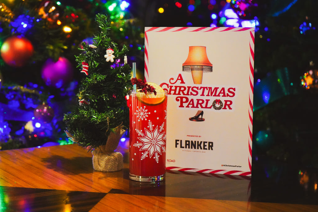 Image pop-up Red holiday cocktail in a snowflake-decorated glass beside a miniature Christmas tree and an A Christmas Parlor sign at Flanker.