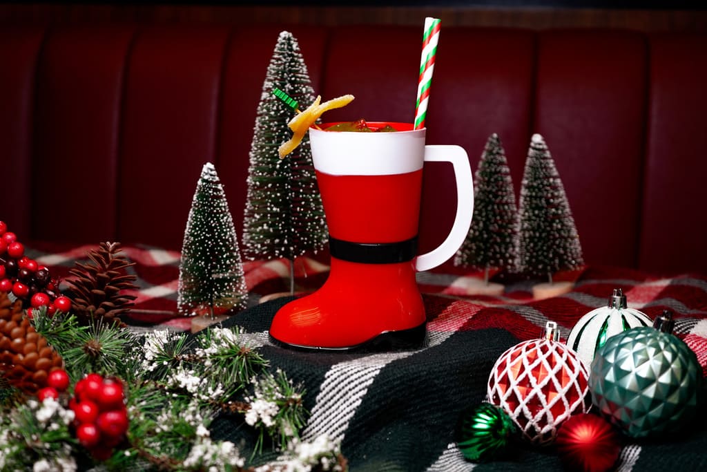 Image pop-up Festive red cocktail served in a Santa boot-shaped mug with a striped straw and citrus garnish, surrounded by Christmas ornaments and miniature snowy trees.