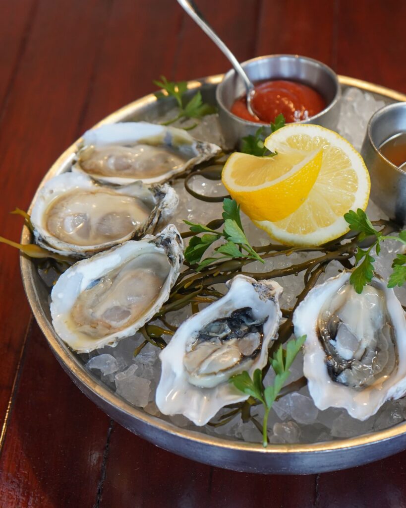 Image pop-up Close-up of fresh oysters on the half shell served over crushed ice with lemon wedges, parsley, and cocktail sauce in a metal bowl.