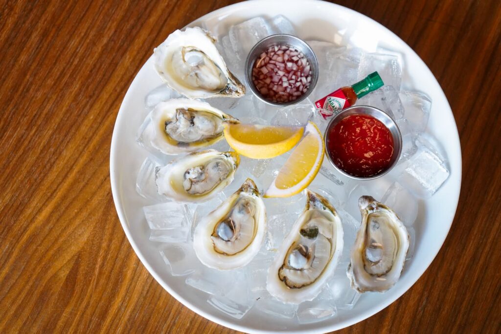 Image pop-up Six fresh oysters on the half shell arranged over ice on a white platter with lemon wedges, cocktail sauce, mignonette sauce, and a small bottle of hot sauce.